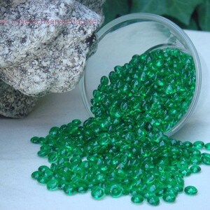 Peut inclure: Un bol en verre rempli de perles de verre vertes se renverse sur une surface blanche. Les perles sont petites et rondes, et elles ont un léger éclat iridescent.