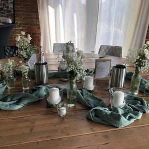 May include: A wooden table set for a special event, featuring white flowers in glass bottles, candles, and teal fabric runners. Stainless steel wine chillers and a framed sign are also present, creating an elegant ambiance.