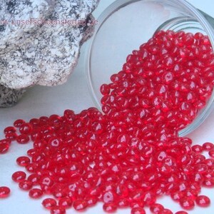 Peut inclure: Un bocal en verre rempli de perles de verre rouge vif. Les perles sont rondes et légèrement aplaties. Le bocal se renverse sur une surface blanche.