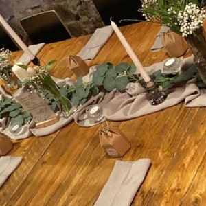 Op de afbeelding: Een rustieke houten tafelsetting met een lange loper van lichtgrijs stof. De tafel is versierd met eucalyptusgroen, witte bloemen en kleine witte kaarsen. Er zijn ook kleine cadeaudoosjes verpakt in bruin papier met touw.