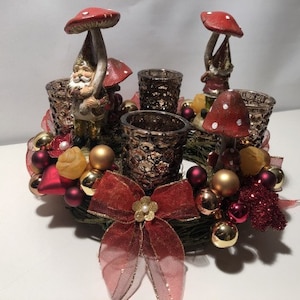 Op de afbeelding: Een rood en gouden kerststuk met een krans, rode en gouden ornamenten en drie glazen kaarshouders met paddenstoelfiguren met kabouters.