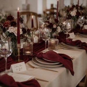 Op de afbeelding: Een lange eettafel gedekt voor een formeel evenement met een crèmekleurig tafelkleed, donkerrode stoffen tafellopers en bijpassende servetten. De tafel is voorzien van wijnglazen, goudkleurig bestek, geribbelde kaarsenhouders en bloemstukken.