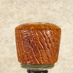 Peut inclure: Un bouchon de bouteille en bois avec un motif sculpté et texturé. Le bouchon est de couleur brun chaud et repose sur une base sombre et ornée. Le motif présente un motif en spirale.
