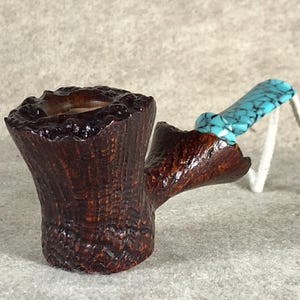 Pode incluir: Um cachimbo de madeira marrom escuro com um bocal turquesa. O cachimbo tem a forma de um tronco de árvore e tem uma tigela no topo.