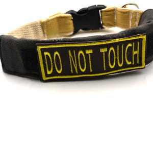 May include: A black dog collar with a yellow and black "DO NOT TOUCH" patch. The collar has a black plastic buckle and a beige interior lining. The collar is made of a durable material.
