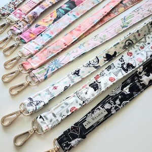 May include: A collection of fabric lanyards featuring various patterns including floral prints, cartoon characters, and Alice in Wonderland motifs. Each lanyard is equipped with a gold-toned metal swivel lobster clasp. One black lanyard features a silver-toned metal tag with the text Ms. Brown.