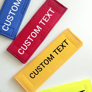 May include: Four fabric straps with the text "CUSTOM TEXT" printed on them. The straps are in blue, red, yellow, and neon yellow.