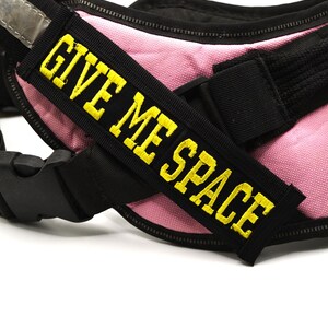 May include: A pink and black dog harness with the yellow text "GIVE ME SPACE". The harness features a reflective silver strip and black buckles. Designed for a dog, it has a zip and adjustable straps.