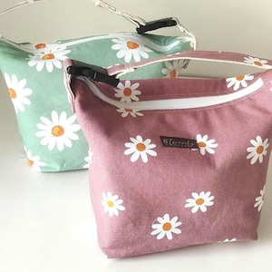 Könnte beinhalten: Zwei Stofftaschen mit Gänseblümchenmustern. Eine Tasche ist hellgrün, die andere mauvefarben. Beide haben weiße Gänseblümchenblüten mit gelben Zentren. Jede Tasche hat einen weißen Reißverschluss und einen Griff. Die mauvefarbene Tasche hat ein kleines schwarzes Etikett mit weißem Text.