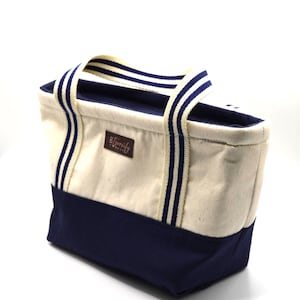 Könnte beinhalten: Eine Canvas-Tragetasche mit cremefarbenem Korpus und marineblauem Boden. Die Tasche hat cremefarbene Griffe mit marineblauen Streifen. Der Markenname "8 Twenty" befindet sich auf einem braunen Lederaufnäher.
