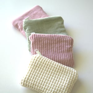 May include: Four small zippered pouches in different colours and textures. The pouches are stacked on top of each other. The top pouch is pink and has a smooth texture. The second pouch is green and has a corduroy texture. The third pouch is pink and has a corduroy texture. The bottom pouch is white and has a waffle texture.
