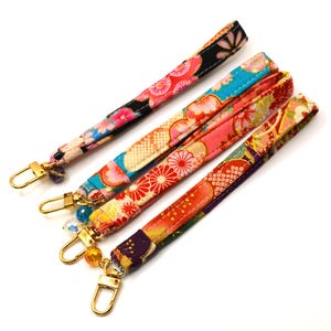 May include: Four fabric wristlet lanyards featuring vibrant, multi-coloured floral and geometric patterns. Each strap is finished with a gold-toned metal lobster clasp and a small decorative bead attached to the base of the hardware.