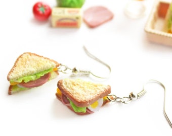 May include: A pair of silver dangle earrings featuring miniature, realistic-looking sandwiches. Each sandwich has two slices of bread, lettuce, ham, and a slice of cheese.