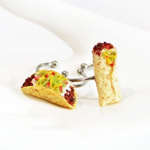 Pode incluir: Dois anéis de comida em miniatura, um com a forma de um taco e o outro com a forma de um burrito. O anel de taco tem uma casca marrom, queijo amarelo, alface verde e molho vermelho. O anel de burrito é marrom e tem queijo amarelo e molho vermelho. Ambos os anéis estão em uma superfície branca.