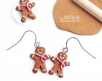 May include: A pair of gingerbread man earrings made from polymer clay. The earrings are brown with white icing and pink ribbon accents. The earrings are shown next to a gingerbread man cookie, a rolling pin, and a small container of butter.
