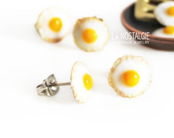 Könnte beinhalten: Vier Paar Ohrringe aus Polymer Clay in Form von Spiegeleiern. Die Ohrringe sind hellbraun mit einem gelben Eigelb in der Mitte. Die Ohrringe werden auf einer weißen Oberfläche mit einer Miniatur-Bratpfanne und einem Spatel im Hintergrund präsentiert.
