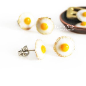 May include: Four pairs of polymer clay earrings shaped like fried eggs. The earrings are a light brown color with a yellow yolk in the center. The earrings are displayed on a white surface with a miniature frying pan and spatula in the background.