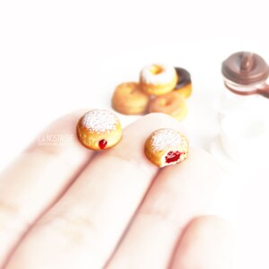 May include: Miniature donuts with powdered sugar and jelly filling, alongside a small coffee pot. Several other donuts are in the background. The image is set against a bright white background. The text "LA NOSTALGIE" is visible.