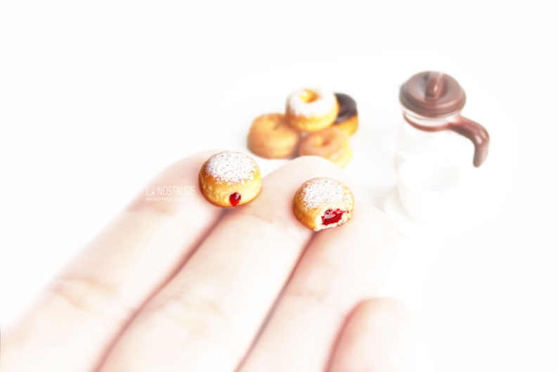 May include: Miniature donuts with powdered sugar and jelly filling, alongside a small coffee pot. Several other donuts are in the background. The image is set against a bright white background. The text "LA NOSTALGIE" is visible.