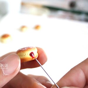 May include: A miniature donut with a light brown exterior and a red filling is being detailed with a needle. The donut is dusted with a white powder. The image includes the text "LA NOSTALGIE HANDMADE JEWELRY".