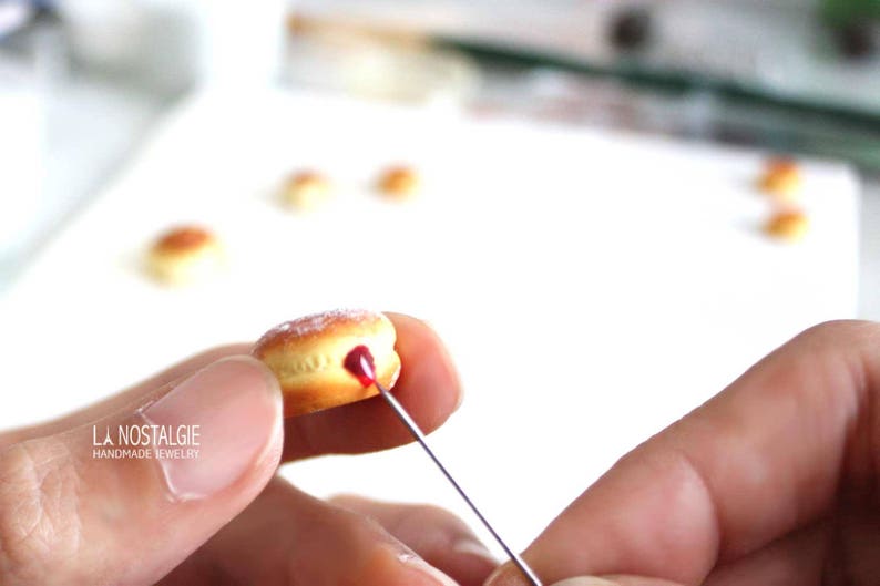 May include: A miniature donut with a light brown exterior and a red filling is being detailed with a needle. The donut is dusted with a white powder. The image includes the text "LA NOSTALGIE HANDMADE JEWELRY".