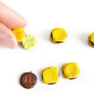 Pode incluir: Um conjunto de brincos de hamb&uacute;rguer em miniatura feitos de argila polim&eacute;rica. Os brincos t&ecirc;m um hamb&uacute;rguer castanho, queijo amarelo e alface verde. Os brincos s&atilde;o exibidos numa superf&iacute;cie branca.