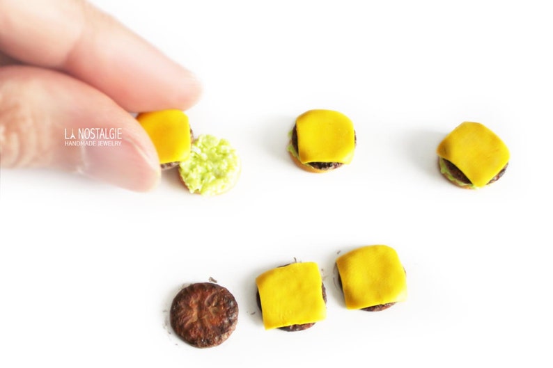 Pode incluir: Um conjunto de brincos de hamb&uacute;rguer em miniatura feitos de argila polim&eacute;rica. Os brincos t&ecirc;m um hamb&uacute;rguer castanho, queijo amarelo e alface verde. Os brincos s&atilde;o exibidos numa superf&iacute;cie branca.
