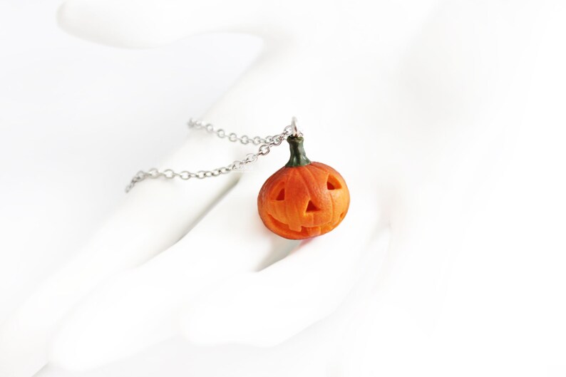 May include: A silver chain necklace with a small orange pumpkin pendant. The pumpkin has a carved jack-o'-lantern face.