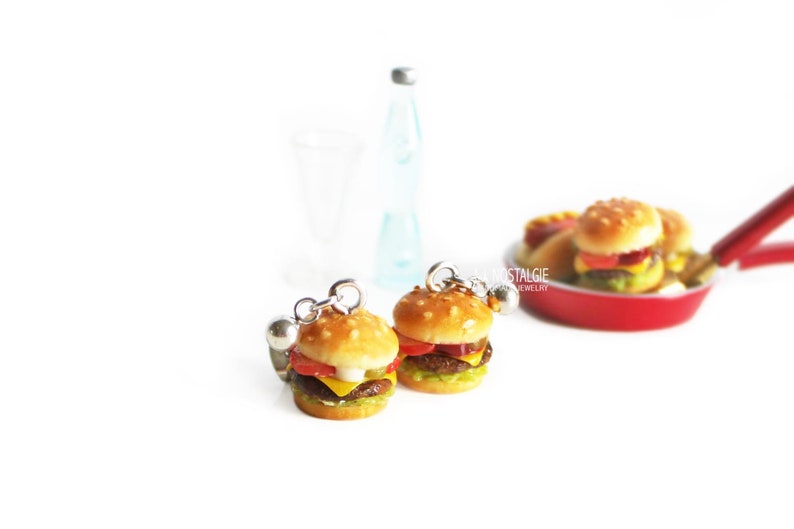 Pode incluir: Um par de brincos pendentes com cheeseburgers em miniatura com alface, tomate, queijo e um p&atilde;o de gergelim. Os brincos s&atilde;o prateados e t&ecirc;m uma pequena bola na parte superior do brinco.