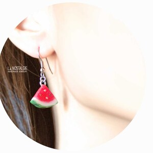 May include: Watermelon slice dangle earrings with silver hooks. The earrings are made of polymer clay and feature a red watermelon slice with black seeds and a green rind.