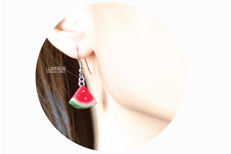 May include: Watermelon slice dangle earrings with silver hooks. The earrings are made of polymer clay and feature a red watermelon slice with black seeds and a green rind.