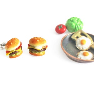 Pode incluir: Um par de brincos de press&atilde;o de prata com hamb&uacute;rgueres em miniatura com alface, tomate, queijo e um hamb&uacute;rguer de carne. Os brincos s&atilde;o exibidos em um fundo branco com uma frigideira em miniatura e uma esp&aacute;tula com dois ovos fritos.