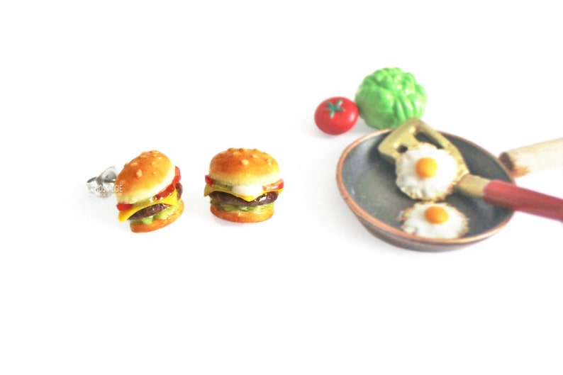 Pode incluir: Um par de brincos de press&atilde;o de prata com hamb&uacute;rgueres em miniatura com alface, tomate, queijo e um hamb&uacute;rguer de carne. Os brincos s&atilde;o exibidos em um fundo branco com uma frigideira em miniatura e uma esp&aacute;tula com dois ovos fritos.