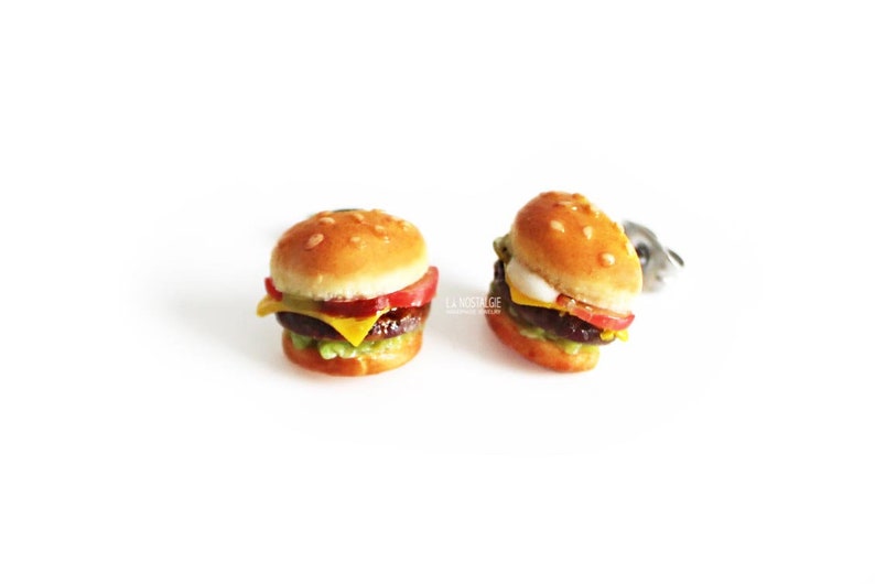 Pode incluir: Um par de brincos de hamb&uacute;rguer em miniatura, cada um com um p&atilde;o de gergelim, alface, tomate, queijo e um hamb&uacute;rguer.