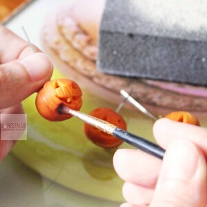 May include: A person is painting a small orange pumpkin with a black paintbrush. The pumpkin has a carved jack-o'-lantern face.