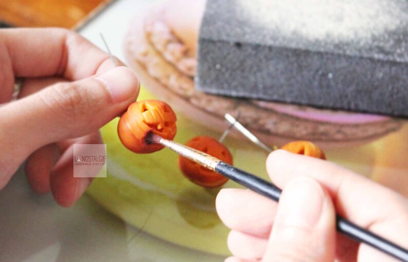 May include: A person is painting a small orange pumpkin with a black paintbrush. The pumpkin has a carved jack-o'-lantern face.