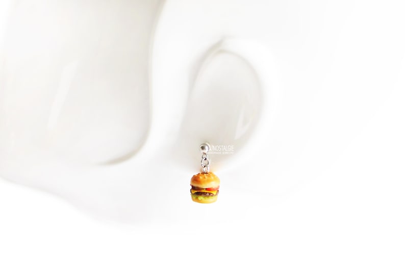 Pode incluir: Um brinco de prata com um mini charme de hamb&uacute;rguer. O hamb&uacute;rguer &eacute; feito de argila polim&eacute;rica e tem um p&atilde;o amarelo, um bife marrom, uma fatia de queijo amarelo e uma folha de alface verde.
