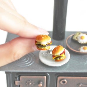 Pode incluir: Dois cheeseburgers em miniatura num prato branco. Os cheeseburgers s&atilde;o feitos de argila polim&eacute;rica e s&atilde;o muito detalhados. Os cheeseburgers est&atilde;o num fog&atilde;o em miniatura.