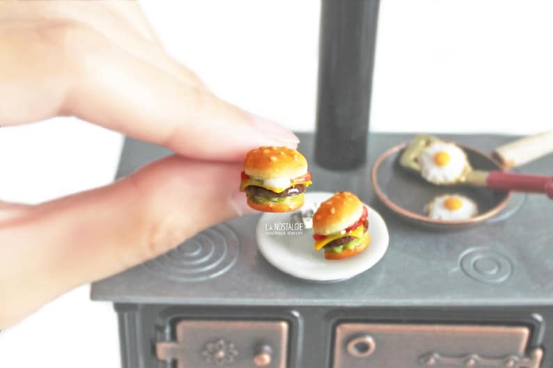 Pode incluir: Dois cheeseburgers em miniatura num prato branco. Os cheeseburgers s&atilde;o feitos de argila polim&eacute;rica e s&atilde;o muito detalhados. Os cheeseburgers est&atilde;o num fog&atilde;o em miniatura.
