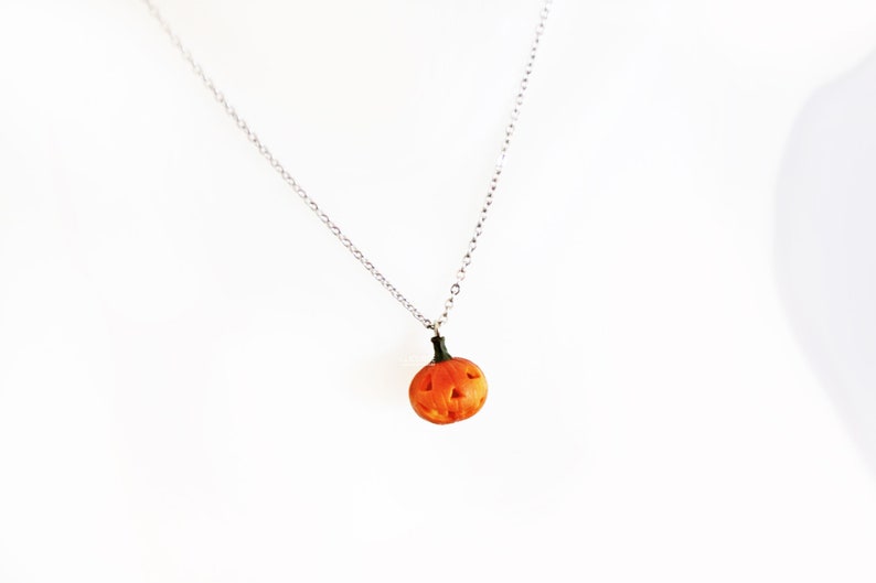 May include: A silver chain necklace with a small orange pumpkin pendant. The pumpkin has a carved face with a black stem.