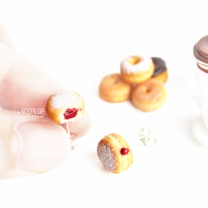 May include: Miniature donut earrings with a bite taken out, revealing a red filling, and dusted with powdered sugar. Other donuts and a coffee pot are in the background. The earrings are handmade jewelry.