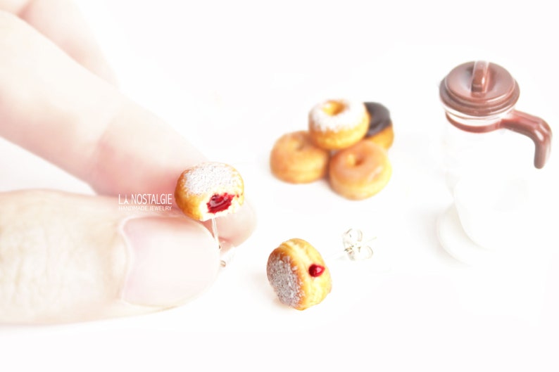 May include: Miniature donut earrings with a bite taken out, revealing a red filling, and dusted with powdered sugar. Other donuts and a coffee pot are in the background. The earrings are handmade jewelry.