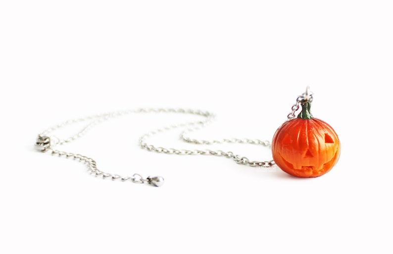 May include: A silver chain necklace with a small orange pumpkin pendant. The pumpkin has a carved jack-o'-lantern face.