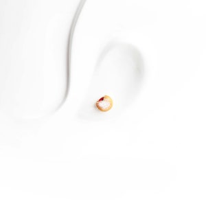 May include: A single stud earring, featuring a small, round design with a light brown base, a red center, and a dusting of white. The earring is set against a white background, highlighting its delicate details.