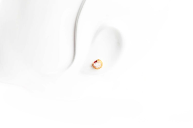 May include: A single stud earring, featuring a small, round design with a light brown base, a red center, and a dusting of white. The earring is set against a white background, highlighting its delicate details.
