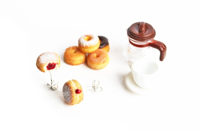 May include: Donut-themed jewelry set featuring two donut earrings, a small coffee pot, and a cup and saucer. The donuts have various toppings, including powdered sugar and glaze. One earring has a bite taken out, revealing a red filling.