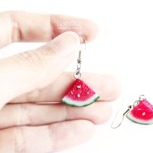 May include: A pair of silver dangle earrings with watermelon slice charms. The watermelon slices are red with black seeds and green rind.