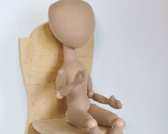 Op de afbeelding: Een beige stoffen pop met scharnierende ledematen zit op een houten stoel. De pop heeft een eenvoudig, rond hoofd en geen gezichtskenmerken. De stoel is gemaakt van licht hout en heeft een eenvoudig, gebogen ontwerp.