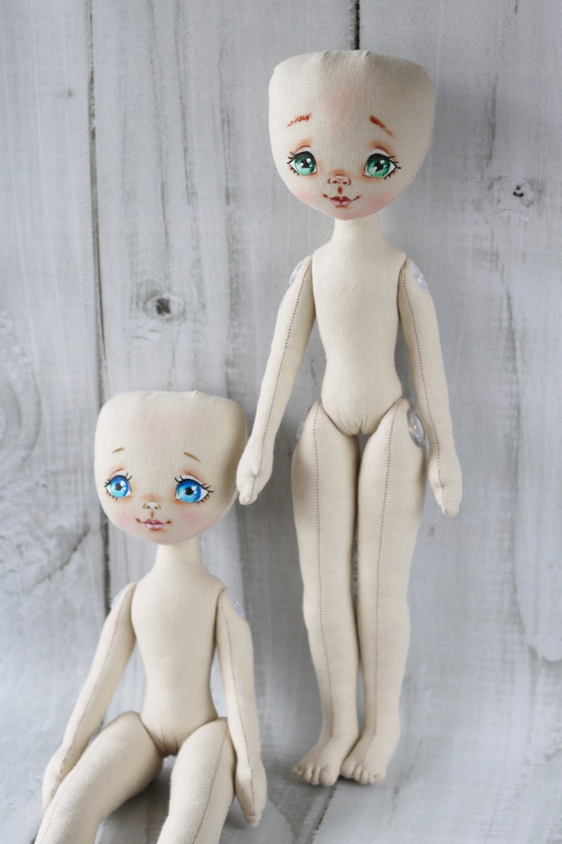 May include: Two handmade fabric dolls with painted faces and blue eyes. The dolls are unclothed and have sewn details. The doll on the left is sitting and the doll on the right is standing.