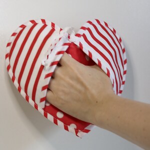May include: A red and white striped heart-shaped pot holder with white polka dots on the inside. The pot holder is made of fabric and has a white string attached to it.